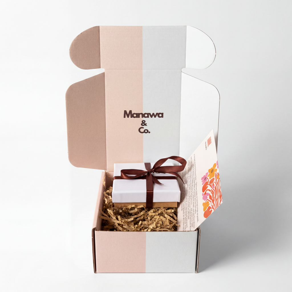 An open Manawa & Co. gift box with a pastel pink, white, and gray color-blocked design, containing a small white gift box tied with a dark brown ribbon on a bed of brown crinkle paper.
