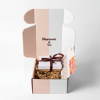 An open Manawa & Co. gift box with a pastel pink, white, and gray color-blocked design, containing a small white gift box tied with a dark brown ribbon on a bed of brown crinkle paper.